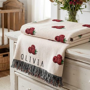 May include: A cream-colored woven throw blanket featuring a repeating pattern of red raspberries and green leaves. The blanket includes the name OLIVIA printed in black capital letters and is finished with a fringed edge along the bottom hem.