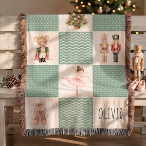 May include: A woven throw blanket featuring a grid pattern with alternating squares of sage green with white polka dots, wavy white lines, and illustrations of a mouse in a crown, a ballerina, nutcrackers, and the name Olivia in black text.