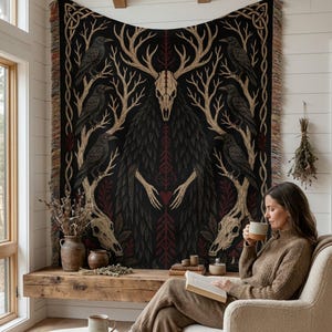 May include: A woven tapestry featuring a central figure with a deer skull head and antlers, surrounded by black ravens perched on bare branches. The design includes a red heart at the center, additional skulls at the bottom, and fringed edges.