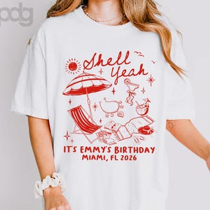 May include: A white t-shirt featuring a red graphic print of beach items including an umbrella, a lounge chair, a cocktail, a seashell necklace, and a towel. The text reads Shell Yeah, IT'S EMMY'S BIRTHDAY MIAMI, FL 2026 in a red font.