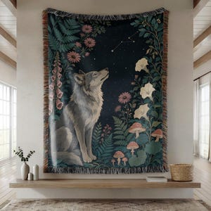 May include: A woven tapestry blanket featuring a grey wolf sitting in a dark forest at night, surrounded by pink flowers, green ferns, white morning glory blossoms, and small mushrooms under a starry sky with a constellation, finished with fringe.