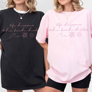 Puede incluir: Dos camisetas holgadas, una negra y otra rosa, con la frase "life happens the beach helps" en escritura rosa. El dise&ntilde;o incluye una ola, una tortuga y un sol. Las camisetas son informales y c&oacute;modas.