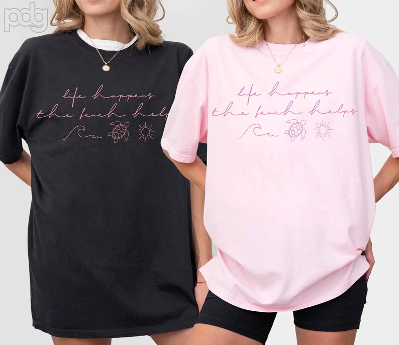 Puede incluir: Dos camisetas holgadas, una negra y otra rosa, con la frase "life happens the beach helps" en escritura rosa. El dise&ntilde;o incluye una ola, una tortuga y un sol. Las camisetas son informales y c&oacute;modas.