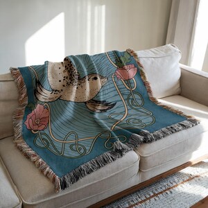 Peut inclure: Une couverture turquoise avec un motif de phoque et des fleurs roses. La couverture a une bordure &agrave; franges et est drap&eacute;e sur un canap&eacute; beige. Le phoque est brun clair avec des taches fonc&eacute;es et les fleurs sont roses. Dimensions: environ 150 cm x 130 cm.