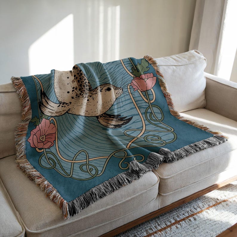 Peut inclure: Une couverture turquoise avec un motif de phoque et des fleurs roses. La couverture a une bordure &agrave; franges et est drap&eacute;e sur un canap&eacute; beige. Le phoque est brun clair avec des taches fonc&eacute;es et les fleurs sont roses. Dimensions: environ 150 cm x 130 cm.