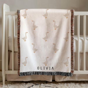 Könnte beinhalten: Eine cremefarbene, gewebte Wohndecke mit einem sich wiederholenden Muster aus hellbraunen Gänsen. Die Decke hat einen Fransenrand mit mehrfarbigen Fäden und der Name OLIVIA ist in schwarzen Großbuchstaben unten in der Mitte aufgedruckt.