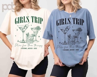 May include: Two women wearing t-shirts with the text "Girls Trip" and a graphic of cocktails. The text below the graphic says "More fun than therapy" and "Cozumel, Mexico - 2025". One shirt is light blue and the other is white.