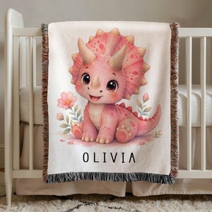 May include: A woven throw blanket featuring a light pink cartoon triceratops dinosaur sitting among small pink flowers. The name OLIVIA is printed in black capital letters below the dinosaur. The blanket has a fringed edge on the top and bottom.