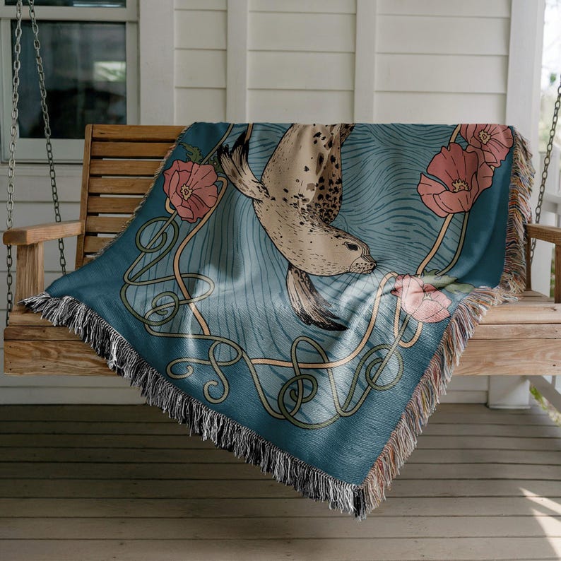 Peut inclure: Une couverture turquoise repr&eacute;sentant un phoque nageant dans l'eau bleue, avec des fleurs roses et des lignes d&eacute;coratives. La couverture a une bordure &agrave; franges et est drap&eacute;e sur une balan&ccedil;oire en bois.