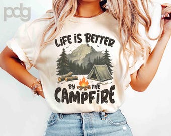 May include: Cream-colored t-shirt with the text "LIFE IS BETTER BY THE CAMPFIRE" in black. The graphic features a mountain scene with a tent, campfire, and trees. The shirt is worn by a person.