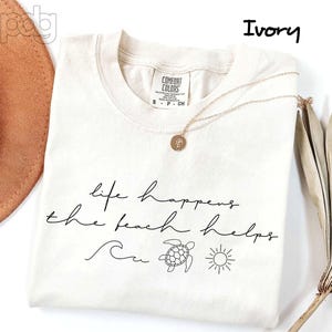 Puede incluir: Camiseta de color marfil con el texto "life happens the beach helps" en escritura negra. La camiseta presenta una ola, una tortuga y un sol. Un collar dorado con un colgante descansa sobre la camiseta.