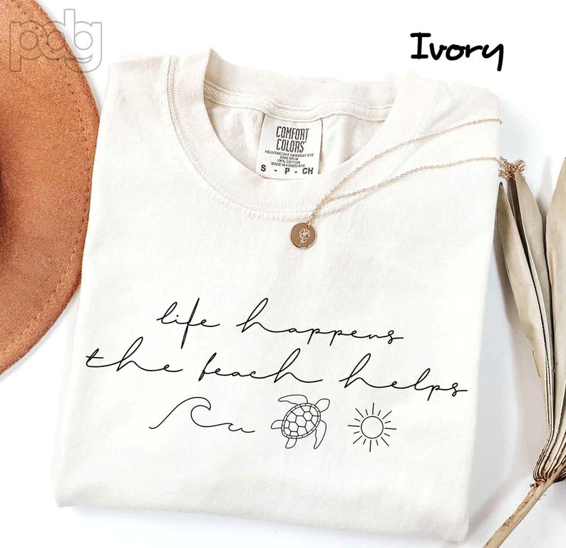 Puede incluir: Camiseta de color marfil con el texto "life happens the beach helps" en escritura negra. La camiseta presenta una ola, una tortuga y un sol. Un collar dorado con un colgante descansa sobre la camiseta.