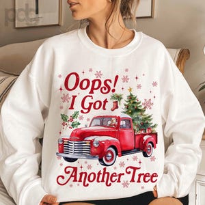 May include: White sweatshirt with a red vintage truck graphic carrying a Christmas tree. The text "Oops! I Got Another Tree" is in red. Snowflakes and holly accents complete the festive design.