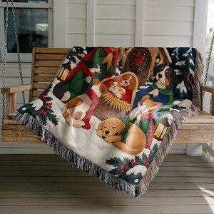 Könnte beinhalten: Eine gewebte Wohndecke mit einer festlichen Winterszene, die mehrere Hunde verschiedener Rassen zeigt, darunter einen Golden Retriever, einen Corgi und einen Welpen in einer Krippe. Das Design umfasst Schnee, Tannenzweige, rote Beeren und Laternen.