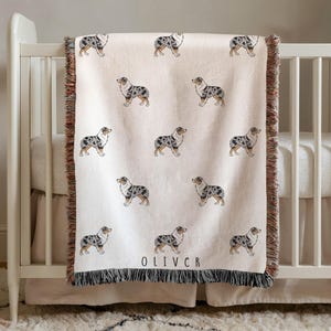 May include: A cream-colored woven throw blanket featuring a repeating pattern of blue merle Australian Shepherd dogs. The blanket has fringed edges on the top and bottom, and the name OLIVER is printed in black capital letters near the bottom edge.