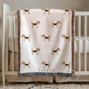May include: A cream-coloured woven throw blanket featuring a repeating pattern of beagle dogs in brown, black, and white. The blanket has fringed edges on the top and bottom, and the name OLIVIA is printed in black text near the bottom edge.