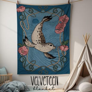 Peut inclure: Une couverture Velveteen turquoise avec une illustration de phoque, des fleurs roses et un cadre de style Art nouveau. La couverture porte les mots "Velveteen" et "blanket".