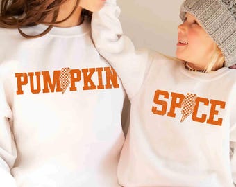 May include: Two white sweatshirts with orange text. One says "PUMPKIN" with a checkered lightning bolt graphic, the other says "SPICE" with a similar graphic. The sweatshirts are worn by two people, one wearing a grey beanie.