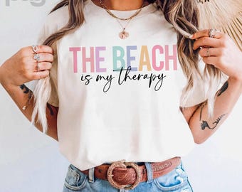 以下が含まれることがあります： パステルカラーと黒い筆記体で「THE BEACH is my therapy」と書かれたクリーム色のTシャツ。青いジーンズと装飾的なバックルが付いた茶色のベルトと組み合わせています。全体的なテーマはビーチとリラックスです。