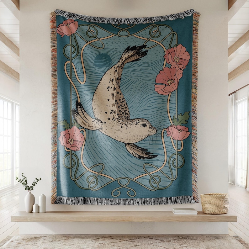 Peut inclure: Une couverture tiss&eacute;e bleu sarcelle avec une illustration de phoque, des fleurs roses et un cadre de style Art nouveau. La couverture a une bordure &agrave; franges et est affich&eacute;e sur un mur.