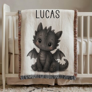 May include: A cream-colored baby blanket with a black dragon illustration and the name "LUCAS" in black letters. The dragon has large eyes and wings. The blanket has a fringed edge and is draped over a white crib.
