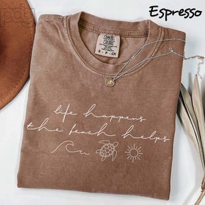 Puede incluir: Camiseta color espresso con la frase "life happens the beach helps" en escritura blanca. La camiseta presenta un gr&aacute;fico de ola, tortuga y sol. Un collar plateado con un colgante descansa sobre la camiseta.