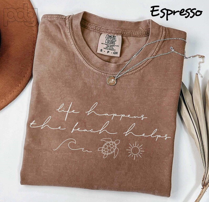 Puede incluir: Camiseta color espresso con la frase "life happens the beach helps" en escritura blanca. La camiseta presenta un gr&aacute;fico de ola, tortuga y sol. Un collar plateado con un colgante descansa sobre la camiseta.