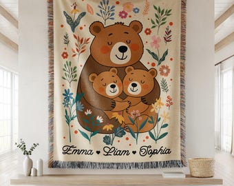May include: Beige woven throw blanket featuring a brown bear embracing two smaller bears, surrounded by colorful flowers and foliage. The names "Emma", "Liam", and "Sophia" are printed at the bottom. The blanket has a fringed edge.