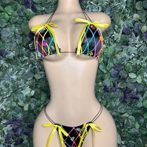May include: A black bikini set with a unique design. The top and bottom feature a black fabric with a colorful grid pattern and small, decorative embellishments. Bright yellow ribbons accent the edges of the top and bottom.