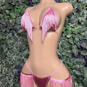 May include: A two-piece bikini set in a light pink satin fabric featuring long, dangling fringe trim along the edges of the triangle top and the sides of the bikini bottoms. The garment is accented with small, clear rhinestones along the seams.