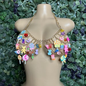 May include: A gold chain bra adorned with colorful candy charms, including lollipops, gummy bears, and donuts. The bra is displayed on a mannequin against a backdrop of green foliage.