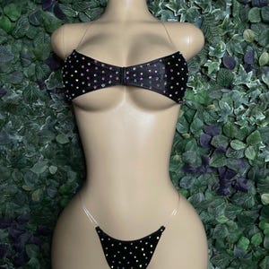 May include: A black two-piece bikini set featuring a front-clasp top and matching bottom, both adorned with scattered iridescent rhinestones. The garment includes thin, clear plastic straps for the top and sides of the bottom piece.