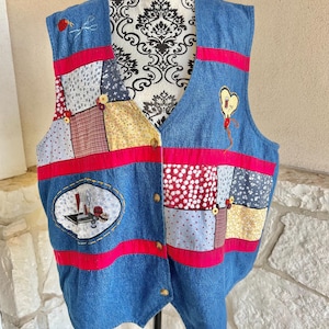 May include: Blue denim vest with patchwork panels in various floral and patterned fabrics. Red ribbon accents and gold-colored buttons adorn the front. Embroidered details include a strawberry and a heart-shaped balloon.