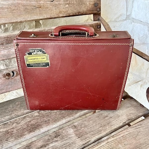 May include: A vintage red leather briefcase with a handle and a metal clasp. The briefcase has white stitching around the edges and a sticker that reads "Guaranteed 5 Years". The briefcase is sitting on a wooden bench.