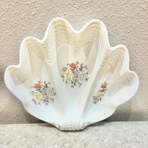 May include: A white, seashell-shaped decorative dish with a scalloped edge. The dish features floral arrangements in yellow, red, and white. The dish is made of ceramic and is suitable for serving or display.