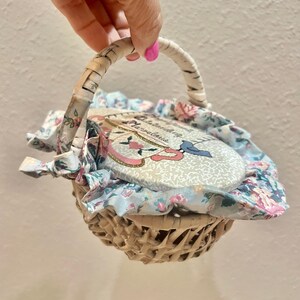 May include: A small, woven basket with a white handle and floral fabric lining. The basket contains a decorative, oval-shaped item with the text "Outstanding & Marvelous" and a bird design. The fabric has a light blue background with pink and white flowers.
