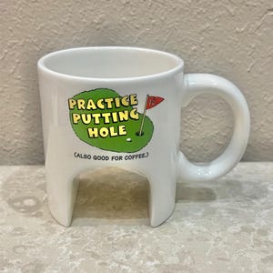 May include: White ceramic coffee mug with a unique design. The mug features the text "PRACTICE PUTTING HOLE" with a golf course graphic. The handle is a large loop. The bottom of the mug has a cut-out design.