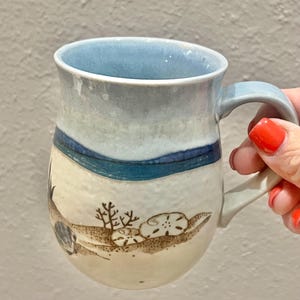 May include: A ceramic mug with a light blue interior and handle. The exterior features a seascape design with a blue sky, a dark blue water line, and a sandy shore with seashells and coral.