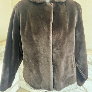 May include: A brown faux fur jacket with a rounded collar and a single button closure. The jacket has long sleeves and a soft, textured appearance. The jacket is displayed on a decorative mannequin.