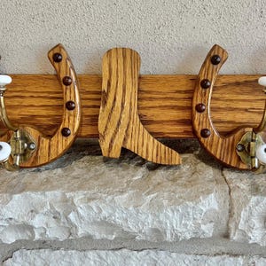May include: Wooden wall-mounted coat rack featuring two horseshoe-shaped hooks and a cowboy boot cutout in the center. The horseshoes have decorative studs and white ceramic knobs. The rack is made of oak wood.