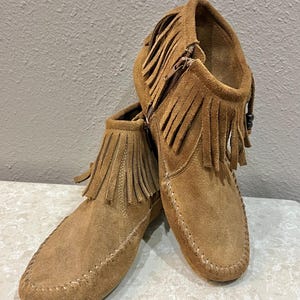 May include: A pair of brown suede moccasin boots with fringe detailing. The boots have a side zipper and are stitched along the seams. The boots are a light brown color and appear to be made of soft suede material.