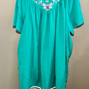 May include: A teal blue short-sleeved dress with a white, pink, and purple floral embroidery design on the neckline. The dress has a white, pink, and purple striped band around the hem.