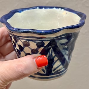 May include: A small, hand-painted ceramic pot with a wavy blue rim. The pot features a white interior and a dark blue and white patterned exterior. The design includes geometric and floral elements. The pot is held by a person.