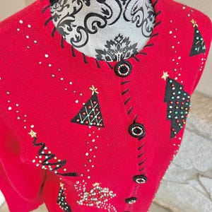May include: A red Christmas sweater with black stitching and black buttons. The sweater features embroidered Christmas trees with colorful bead accents and scattered white and gold embellishments. The neckline is trimmed with black stitching.