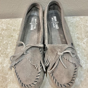 May include: A pair of grey suede moccasins featuring a fringe detail across the vamp, a decorative bow, and black whipstitching along the edges. The interior insoles are printed with the white text Minnetonka Moccasin.