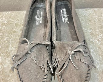 May include: A pair of grey suede moccasins featuring a fringe detail across the vamp, a decorative bow, and black whipstitching along the edges. The interior insoles are printed with the white text Minnetonka Moccasin.