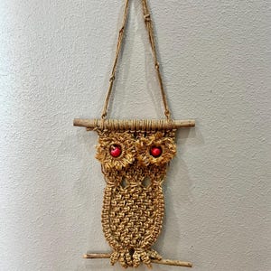 May include: A macrame owl wall hanging. The owl is made of tan-colored rope with red bead eyes. It hangs from a wooden dowel and a rope loop. The owl is perched on a small wooden branch.