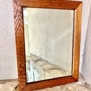 May include: Rectangular mirror with a warm brown wooden frame. The frame has a textured, patterned design on the sides and a smooth finish on the top and bottom. The mirror reflects a light-colored wall and a stone ledge.