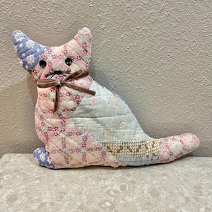 May include: A handmade cat-shaped pillow made from patchwork fabric. The pillow features a variety of floral and star patterns in pink, blue, and cream colors. It has button eyes and a brown ribbon bow.