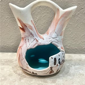 May include: A white ceramic vase with a unique double-spout design and a central opening. The vase features hand-painted details in shades of brown and white, with a turquoise interior. The design includes a depiction of a Native American village.
